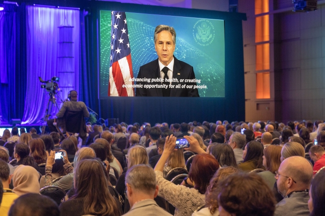 A crowd watches a video of a light-skinned man with gray hair in front of an American flag